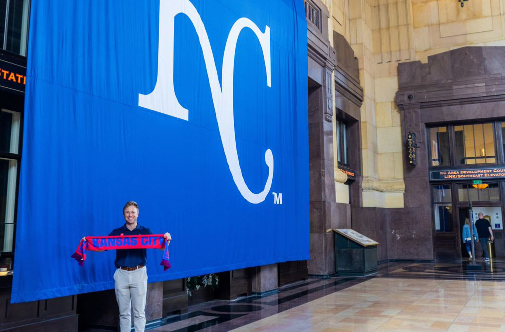 A man stands below a large blue banner that says KC, and he's holding a red banner that says Kansas City in blue letters.
