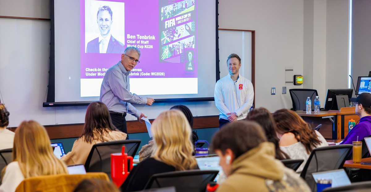 Two men stand in front of a classroom of students in front of a screen that has a purple presentation.