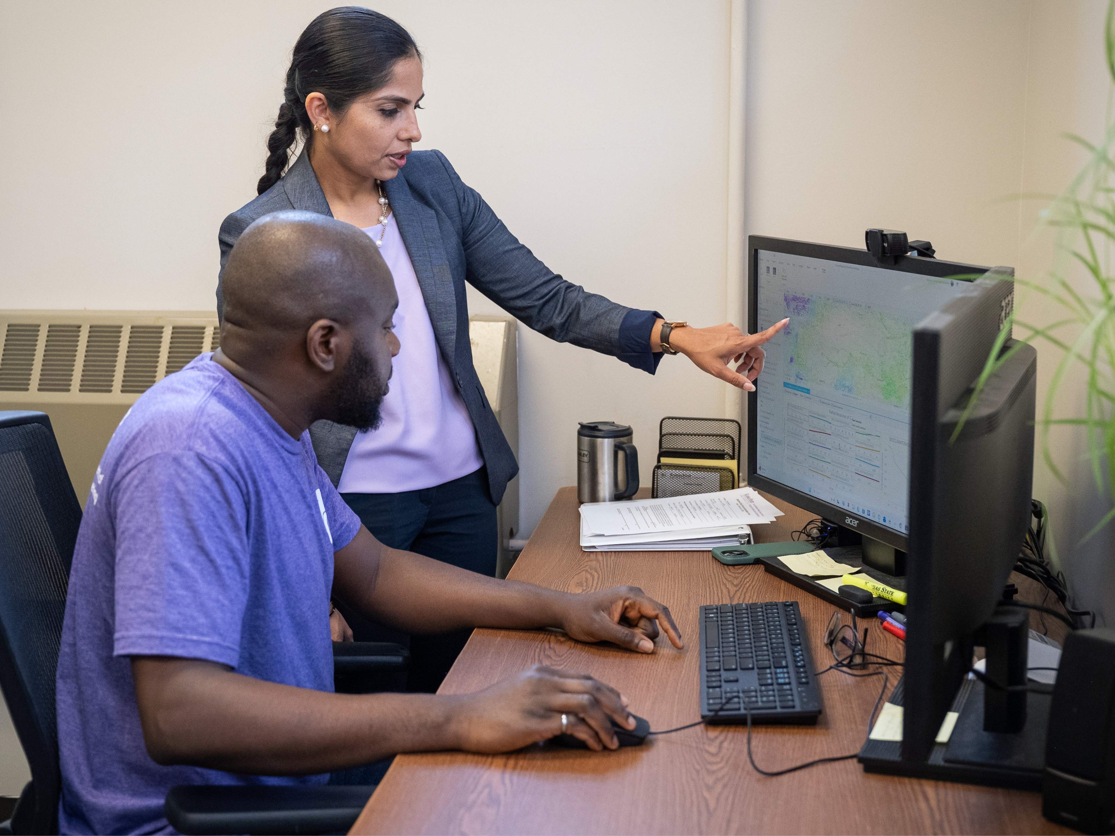 Sharda, wearing a grey blazer and purple top, points at a computer screen. Sitting at the desk is a student wearing a purple t-shirt, who is navigating the mouse on the computer.