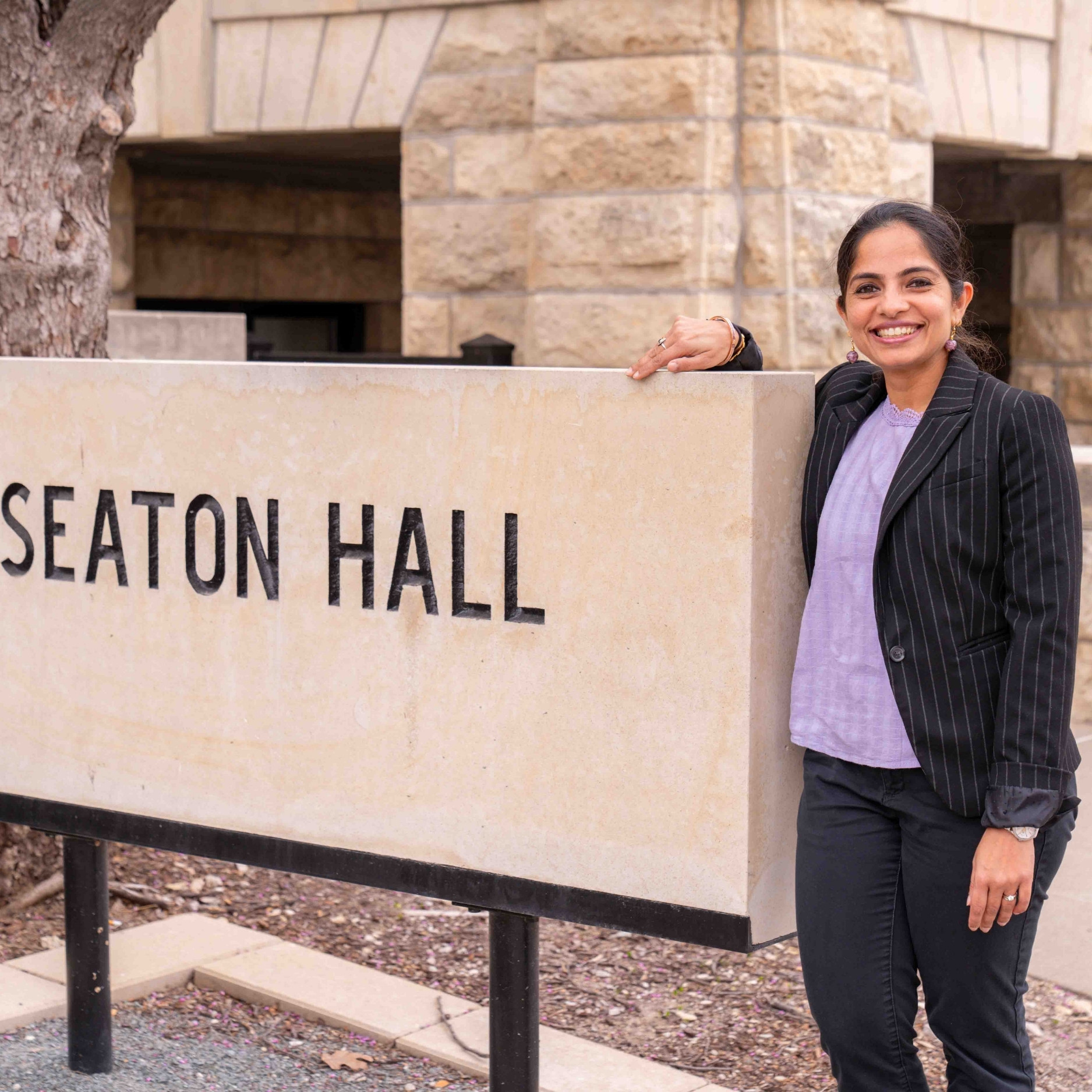 Vaishali Sharda, wearing a lavender blouse and black striped blazer, smiles and poses up against a sign reading 