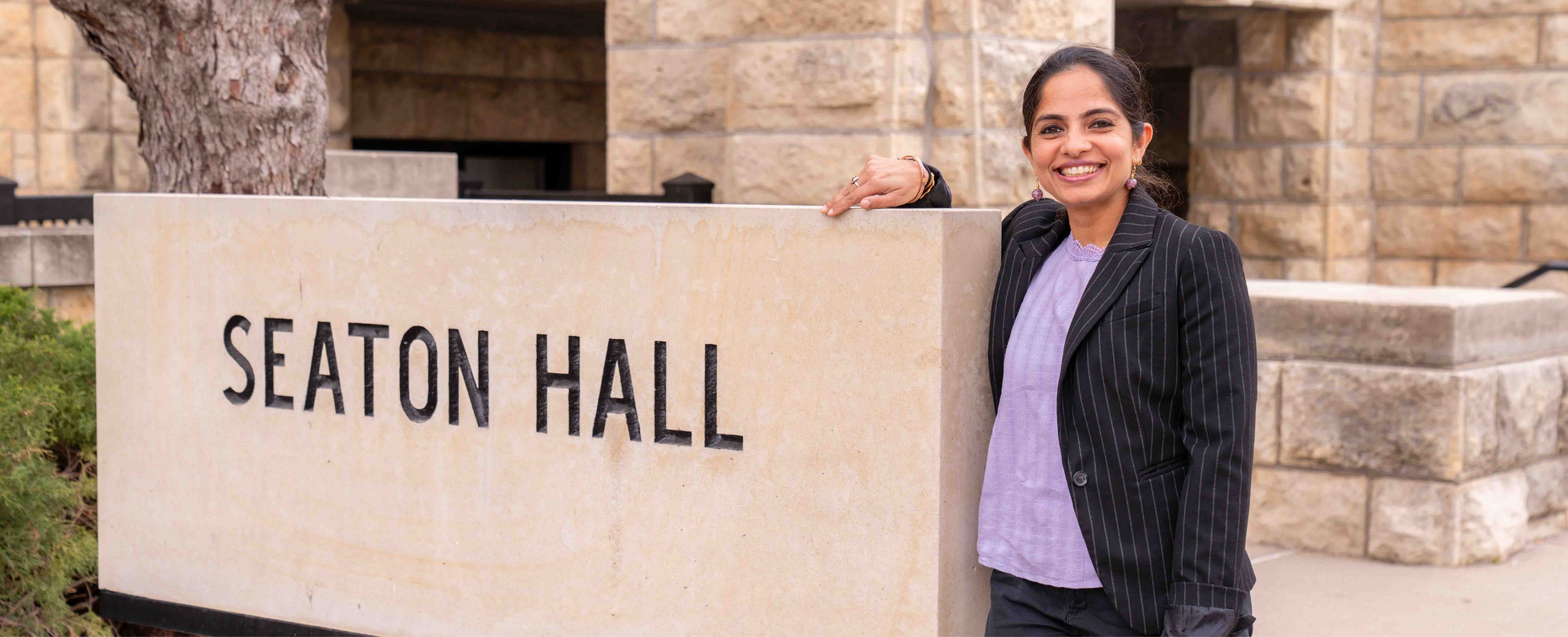 Vaishali Sharda, wearing a lavender blouse and black striped blazer, smiles and poses up against a sign reading "Seaton Hall."