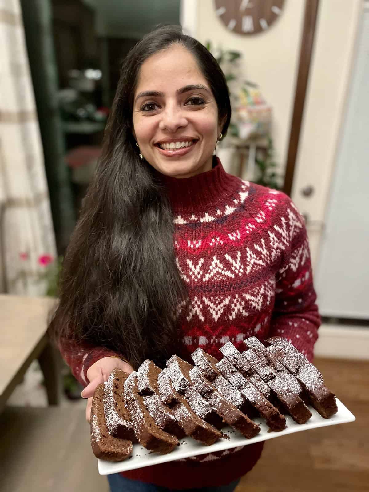 Sharda, wearing a red Christmas sweater, holds a white plate with slices chocolate loaf bread on top.