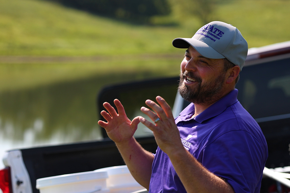 A man in a purple polo and gray hat stands sits in a truck bed and talks to someone out of frame. 