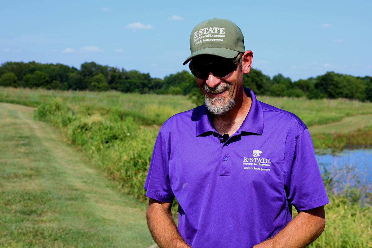 A man in a purple polo and green hat stands in a green field near a pond. 