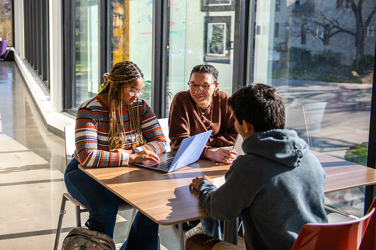 A group of college students meet and discuss at a table inside a brightly lit meeting space.