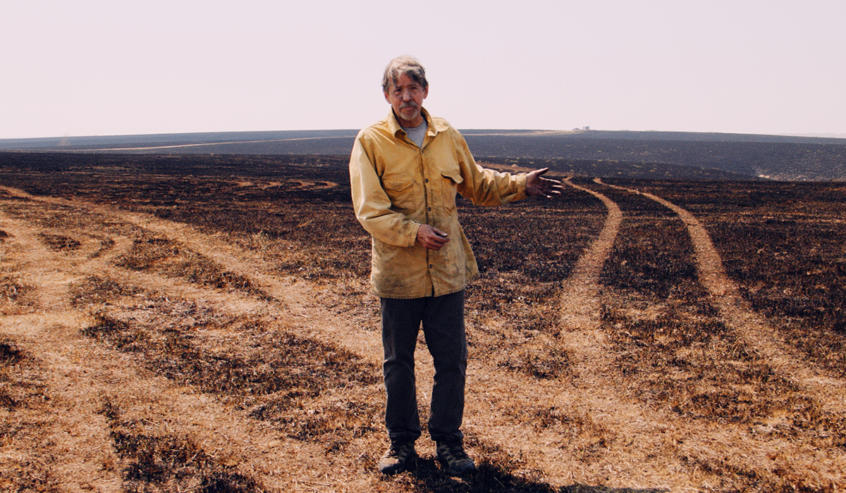 A researcher in a yellow fire jacket stands and explains his research in a burned over section of hilly prairie.