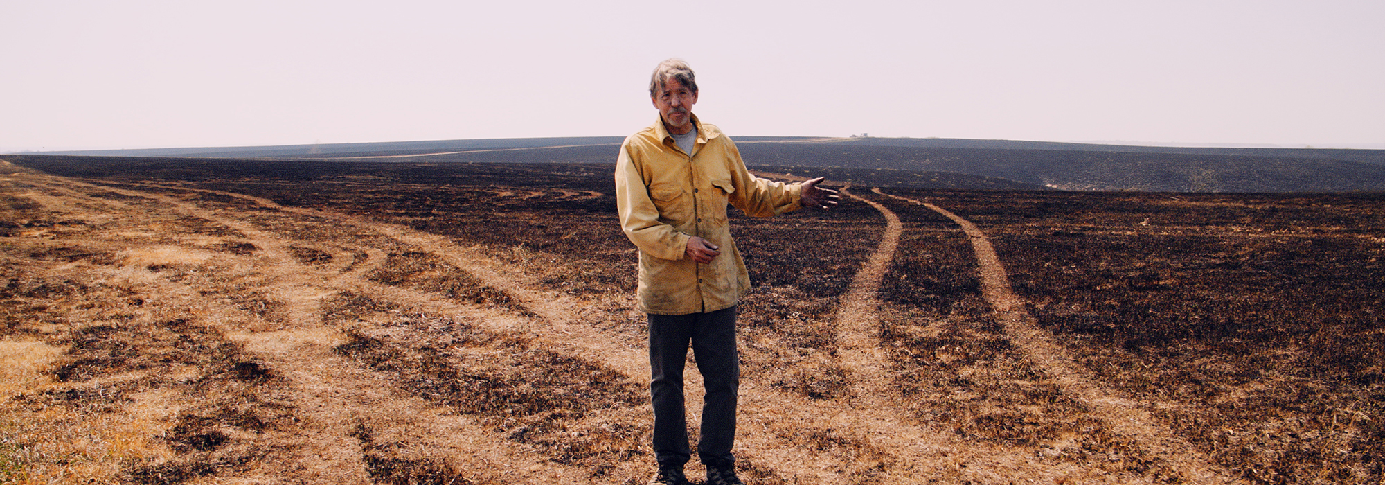A researcher in a yellow fire jacket stands and explains his research in a burned over section of hilly prairie.