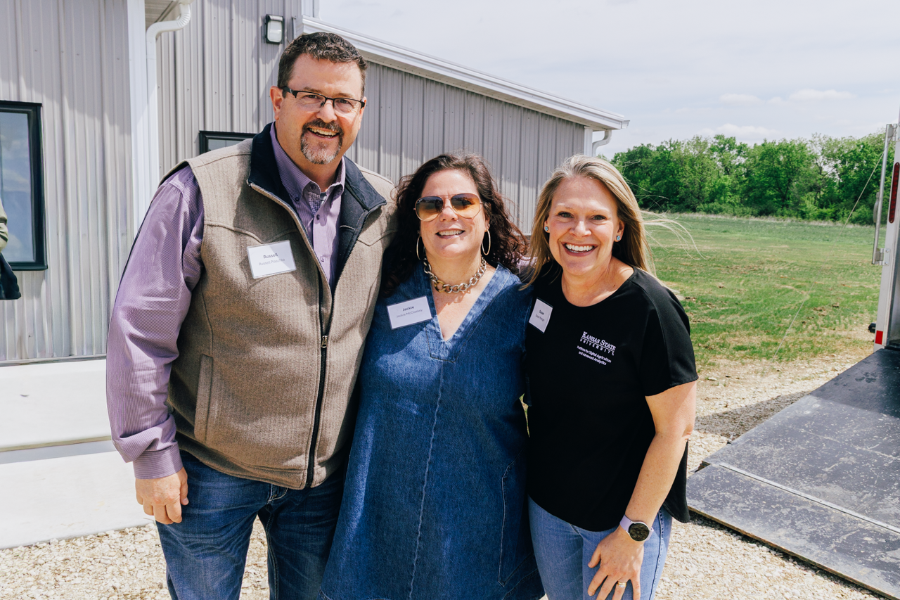 A man and two women stand outside of a building with greenery in the background and smile for a photo