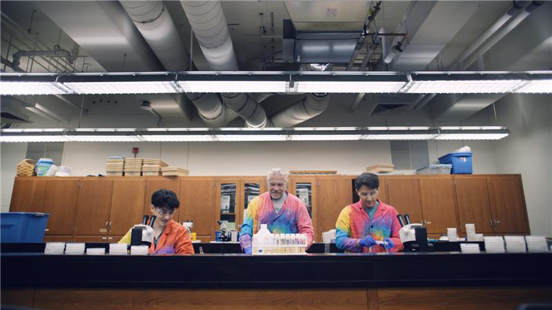 A college professor, with two undergraduate students at his side, works at a bench in a research laboratory. All are wearing tie-dye lab coats.