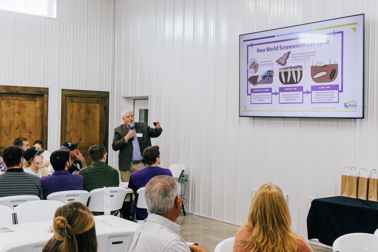 A man presenting at the front of a room gestures toward a PowerPoint slide covering New World screwworm