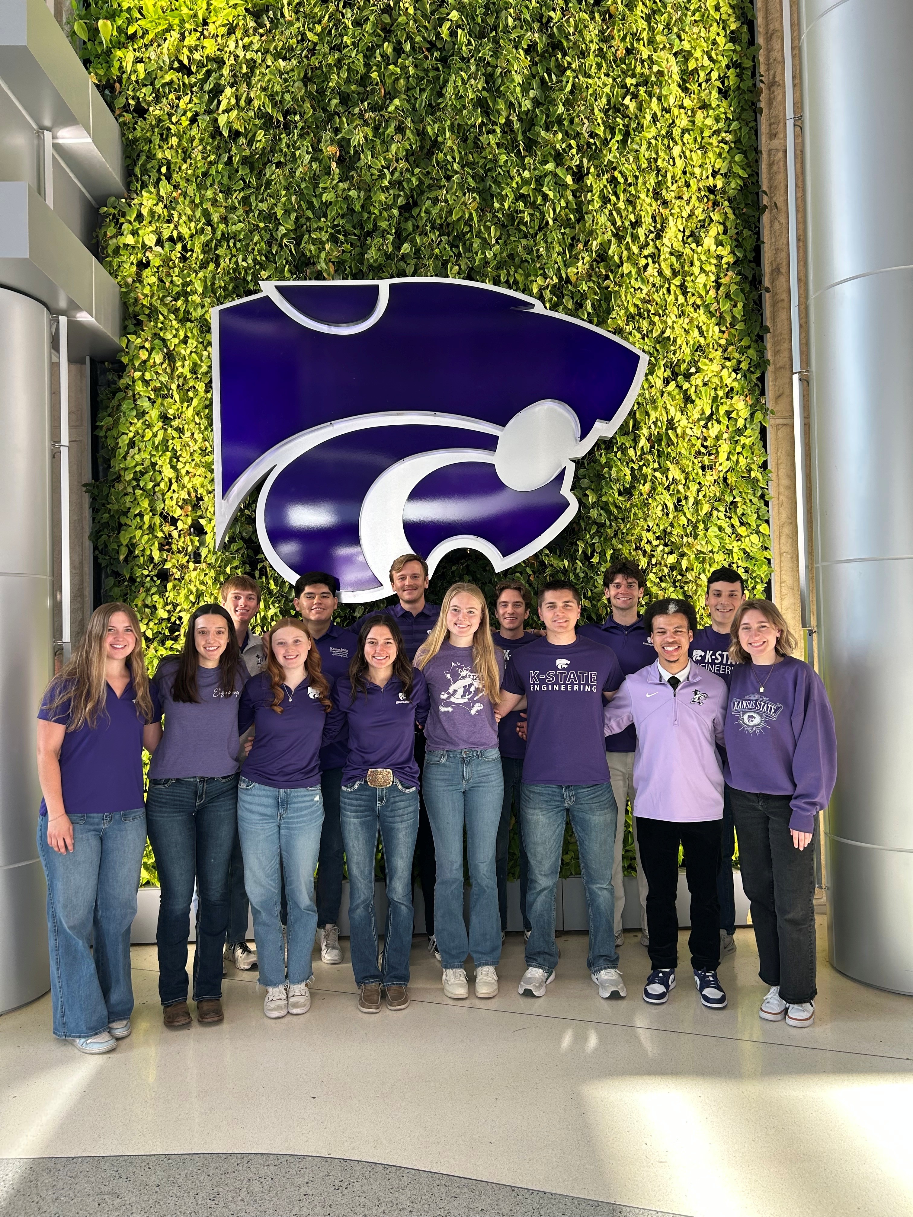 Fourteen students wearing purple shirts stand in front of a wall made of live plants and a large purple Powercat.