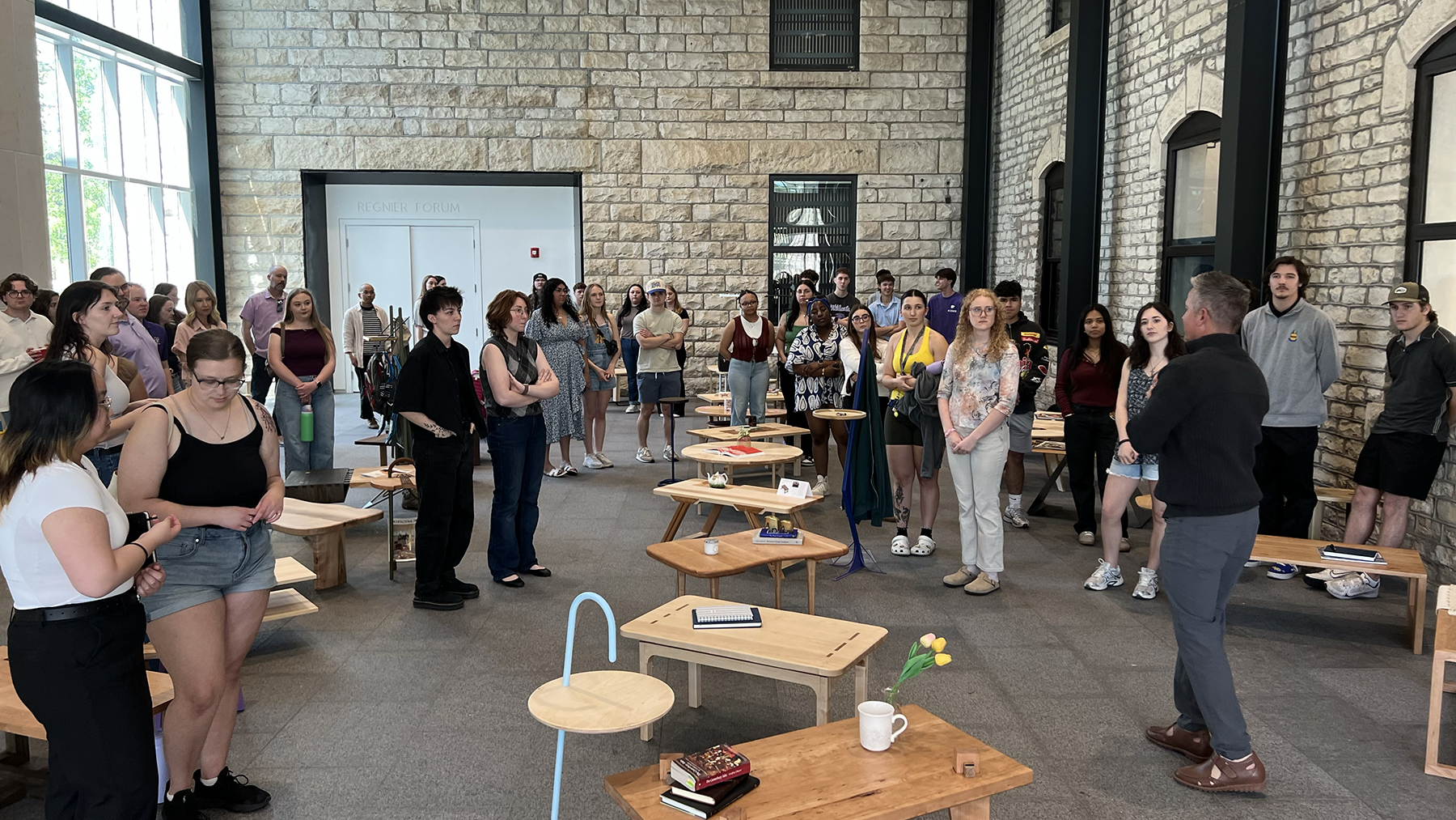 Several people listen to a man speak amid a wooden furniture showcase in an architecture college's lobby area.