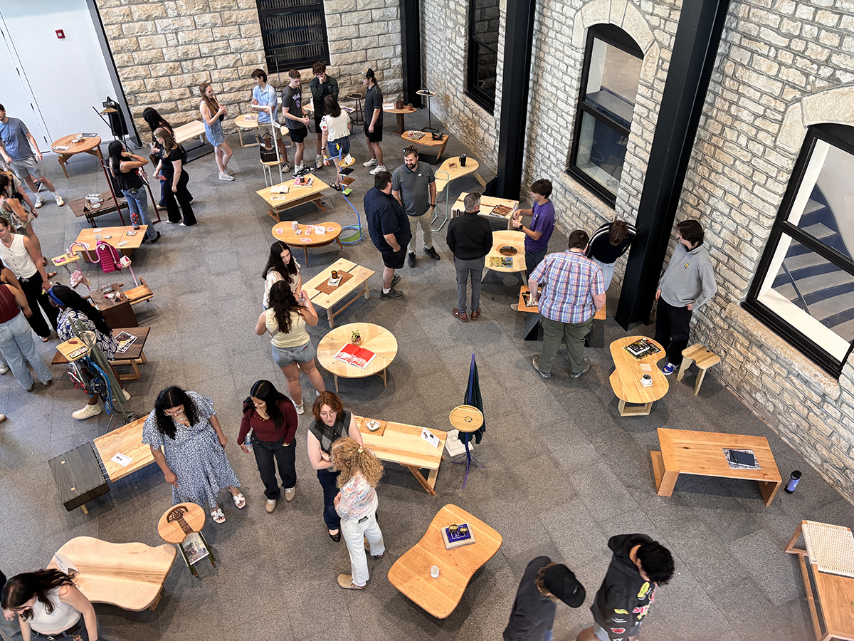 An overhead view shows several people mingling around wooden furniture in a college hall's lobby.
