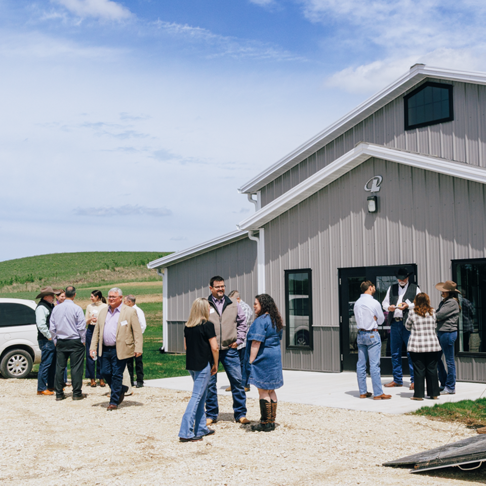 Several groups of people stand outside of a gray building set against a green field and blue sky in the background.