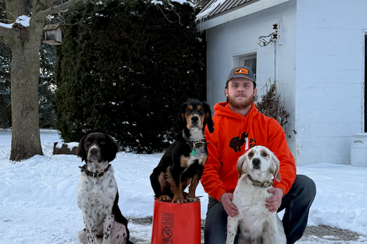 A man wearing an orange sweatshirt and a gray hat kneels outside in the snow, and three dogs sit next to him. 