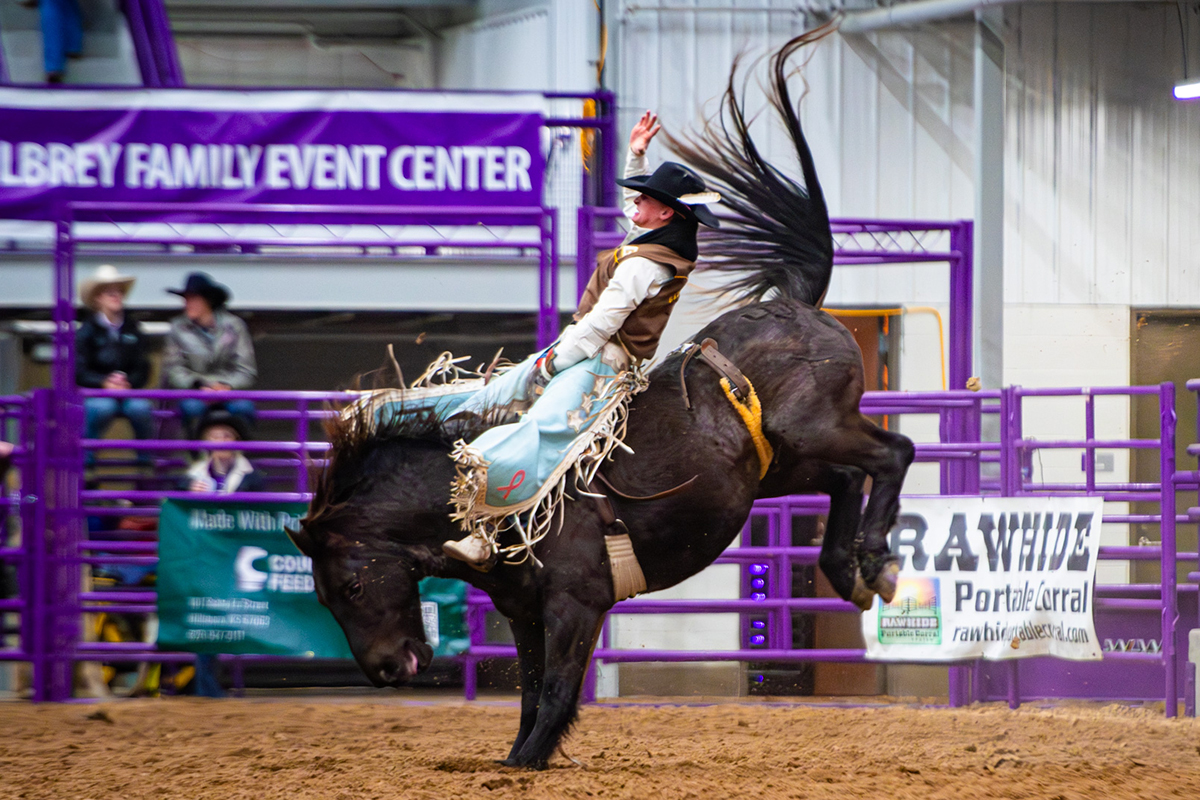 A man in western gear and fancy fringed cowboy pants rides a bucking horse bareback in a rodeo arena.