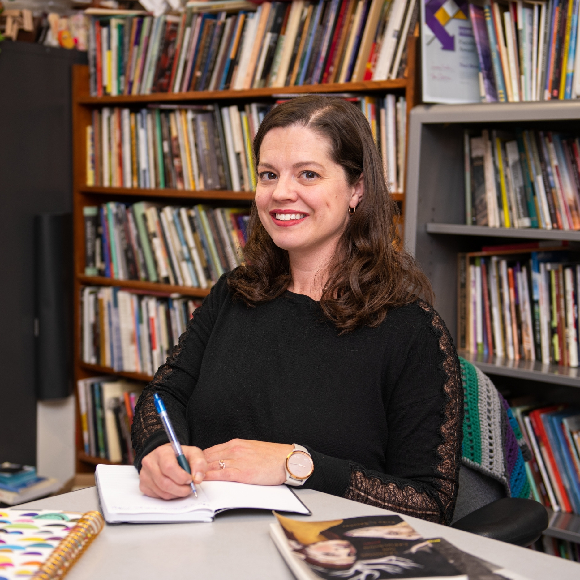 Traci Brimhall sitting at a desk, holding a pen. She is writing in a notebook, with bookshelves behind her.