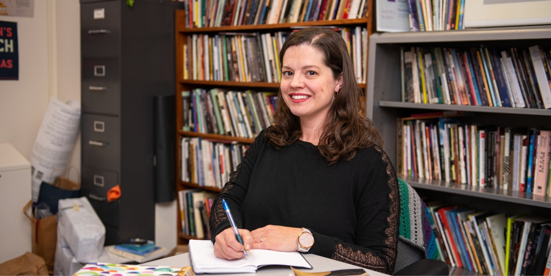 Traci Brimhall sitting at a desk, holding a pen. She is writing in a notebook, with bookshelves behind her.