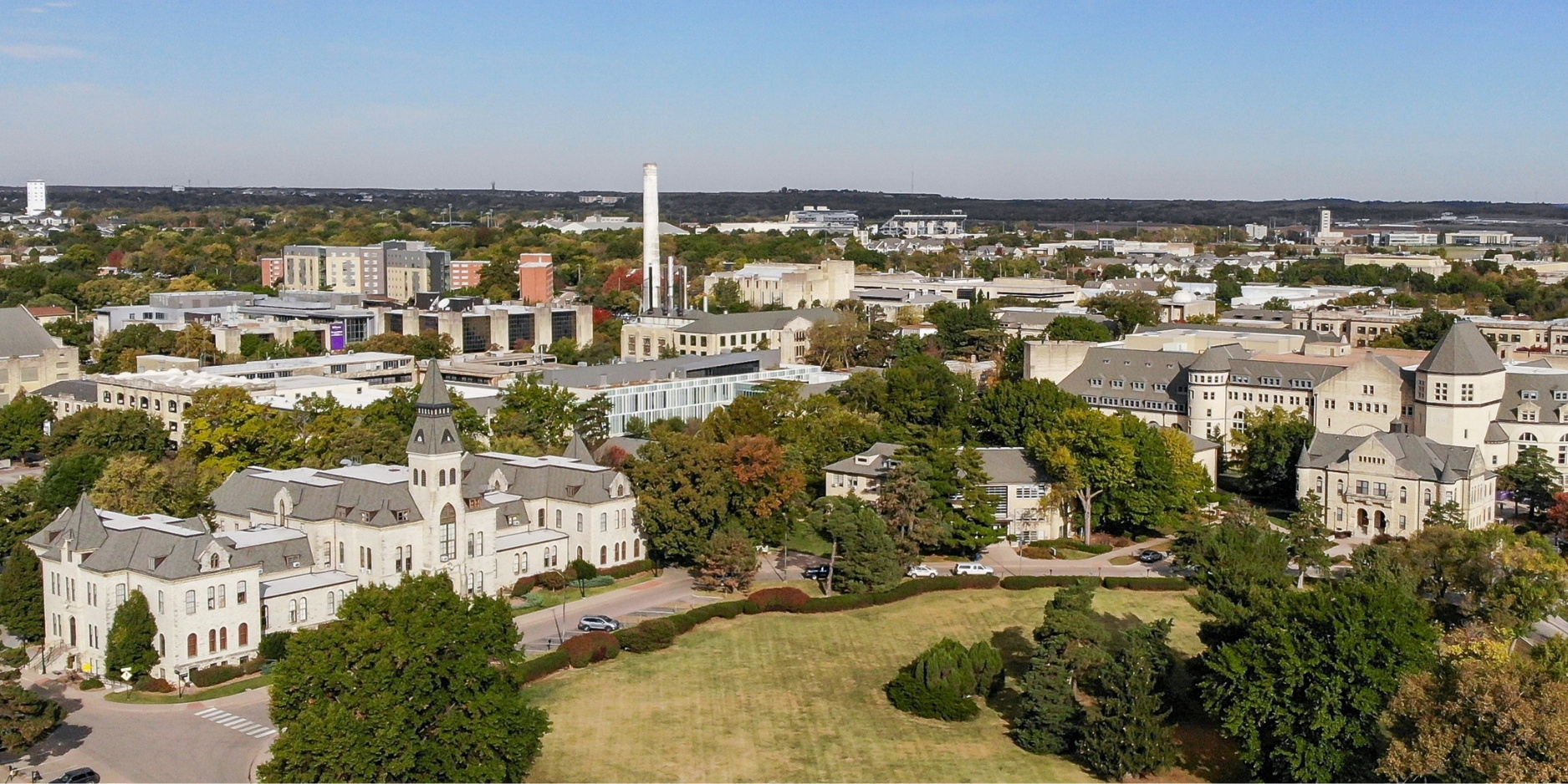 Aerial view of Kansas State University with historic limestone buildings, green lawns and tree-lined streets in sunlight.