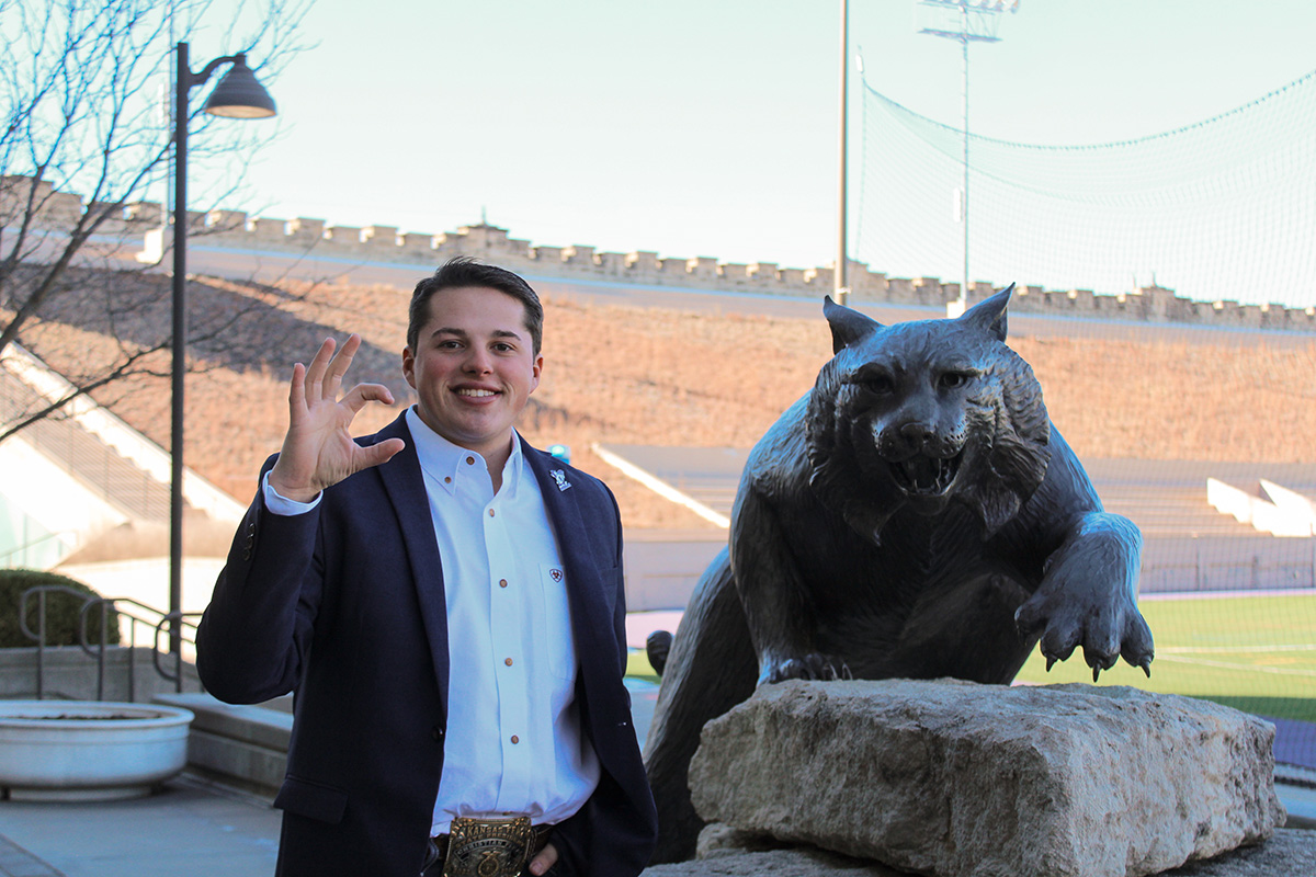 A college student wearing a blazer and a large silver belt buckle smiles next to a metal wildcat statue and does the Kansas State University Wildcat hand-sign. 
