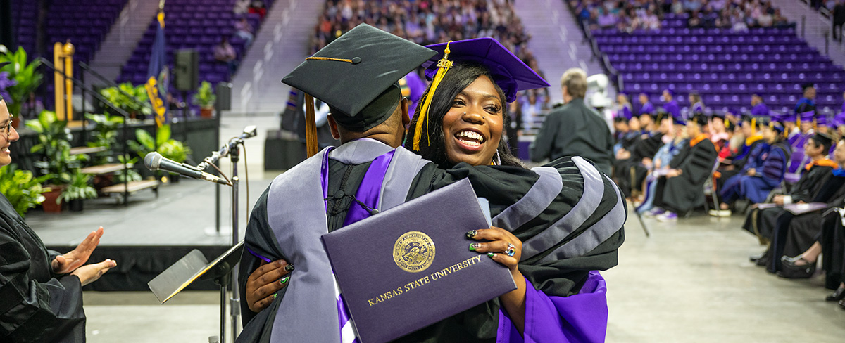 A Black woman wearing purple graduation regalia hugs an administrator while holding her diploma.
