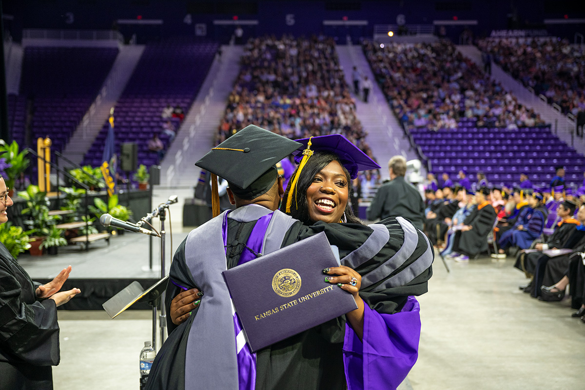 A Black woman wearing purple graduation regalia hugs an administrator while holding her diploma.