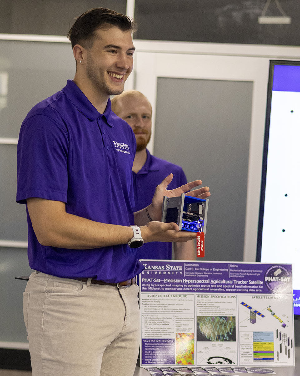 A student holds a 3D model of a miniature satellite as part of a presentation inside a classroom during a college open house.