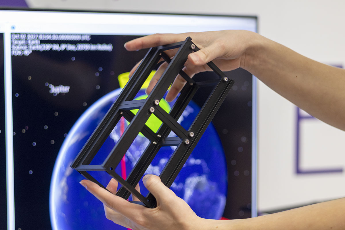 A pair of hands hold a small rectangular frame of a satellite in front of a display with the Earth, as seen from space, on it.