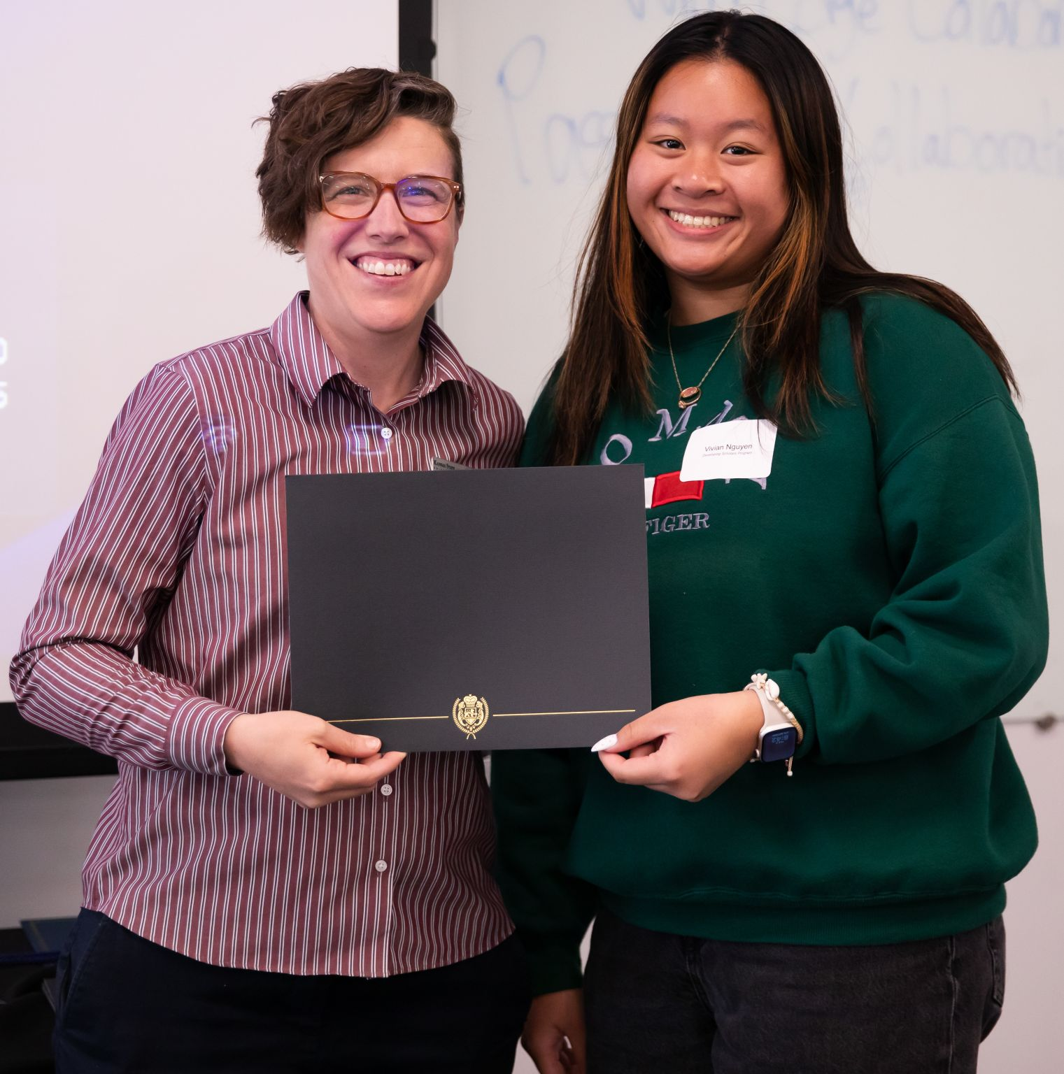 A faculty member in a striped shirt and a student in a green sweater hold an award certificate cover and smile.