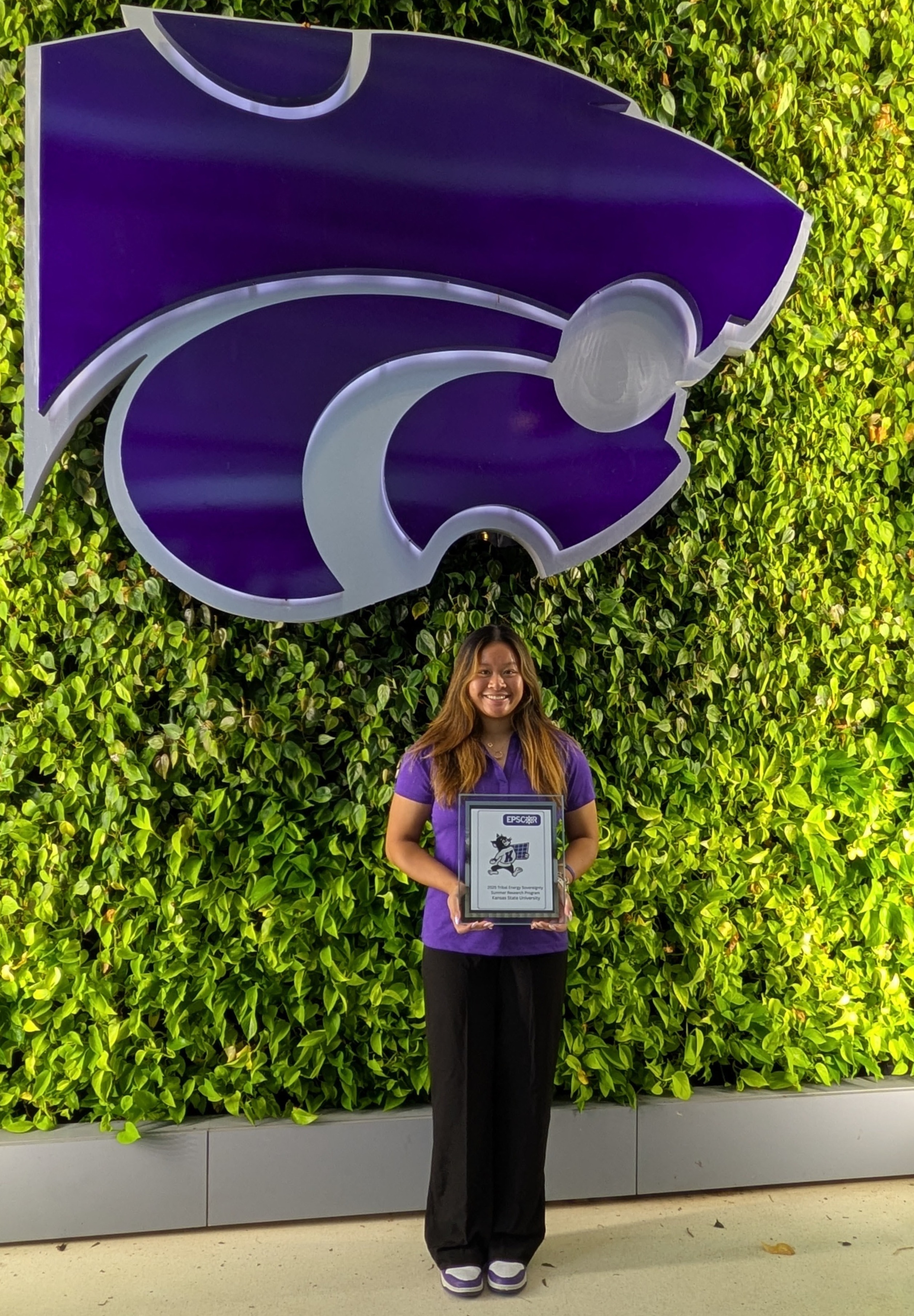 A female student in a purple polo stands in front of a green wall made of plants that also has a purple powercat on it.