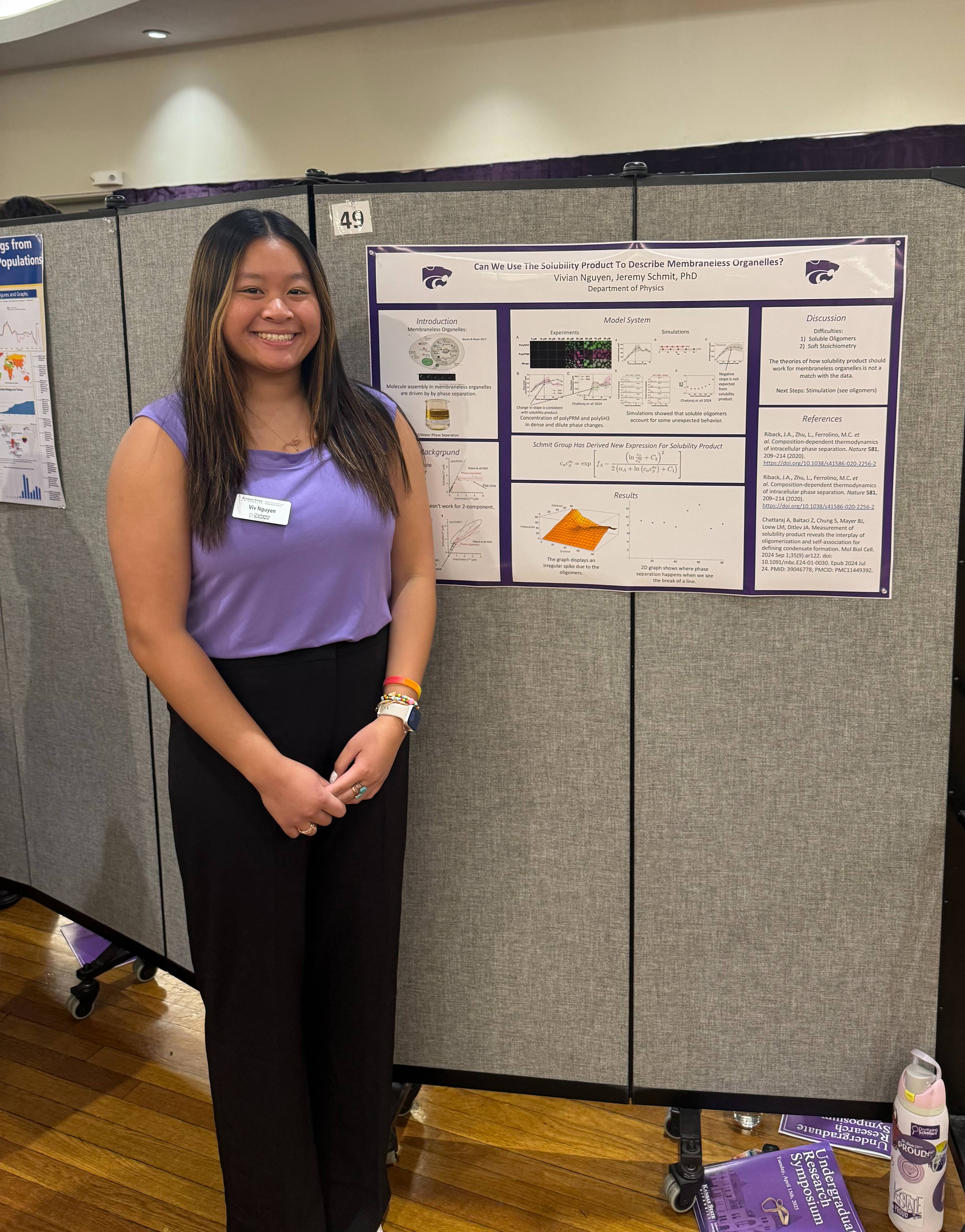 A female student in a purple blouse stands in front of a board that has a research poster posted on it.