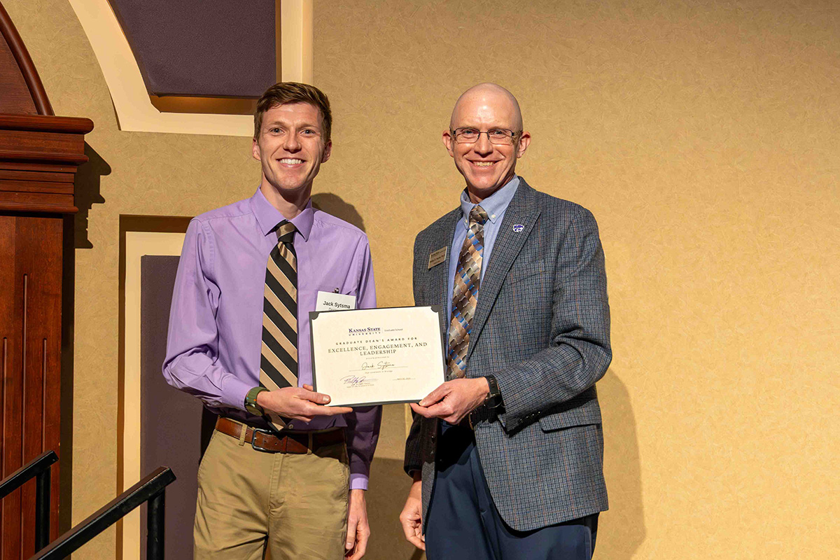 A male graduate student stands next to a college dean who presents him with an award certificate on a stage.