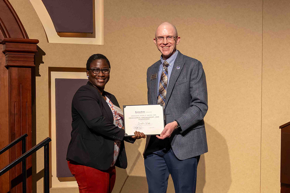 A female graduate student stands next to a college dean who presents her with an award certificate on a stage.