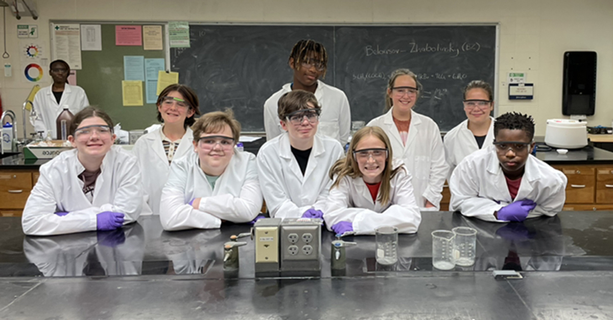 A group of middle school students wearing lab coats, glasses and gloves stand behind a table with glass beakers in a science classroom.