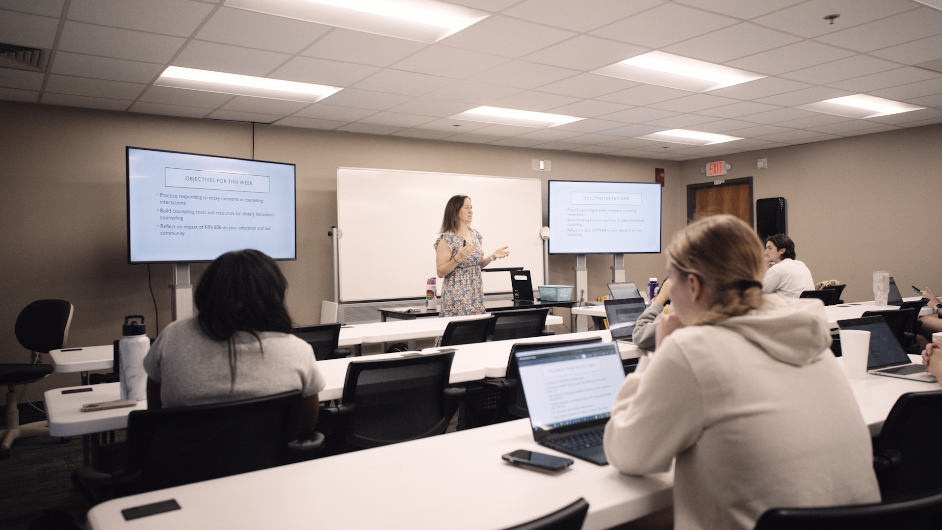 A woman in a floral dress stands at the front of a classroom in front of a whiteboard with screens on either side of her listing the week's objectives. Students sit with laptops at tables in the room.