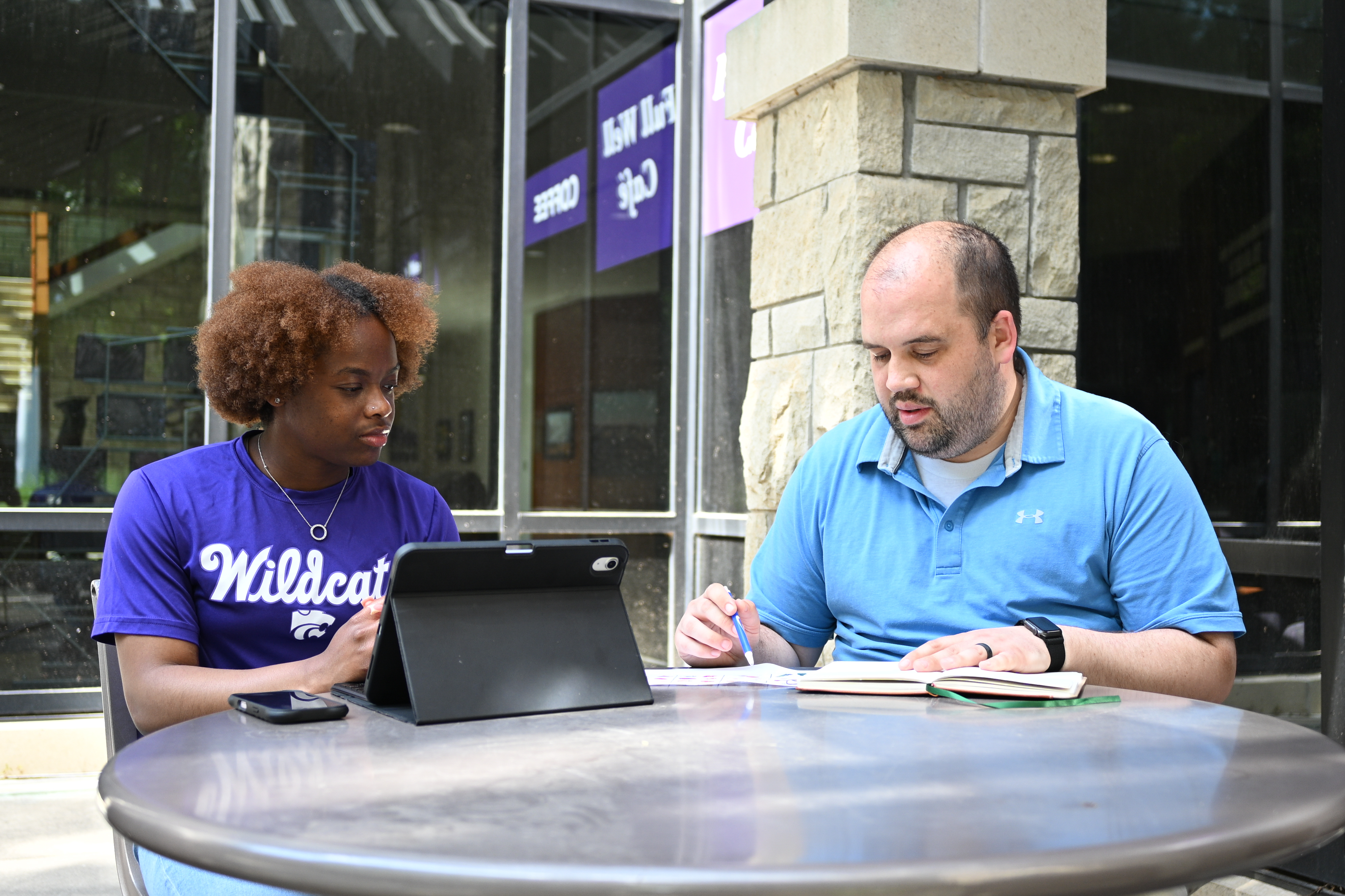 A student in a purple t-shirt sits with an ipad next to a man in a blue polo for a health counseling session at an outdoor table.