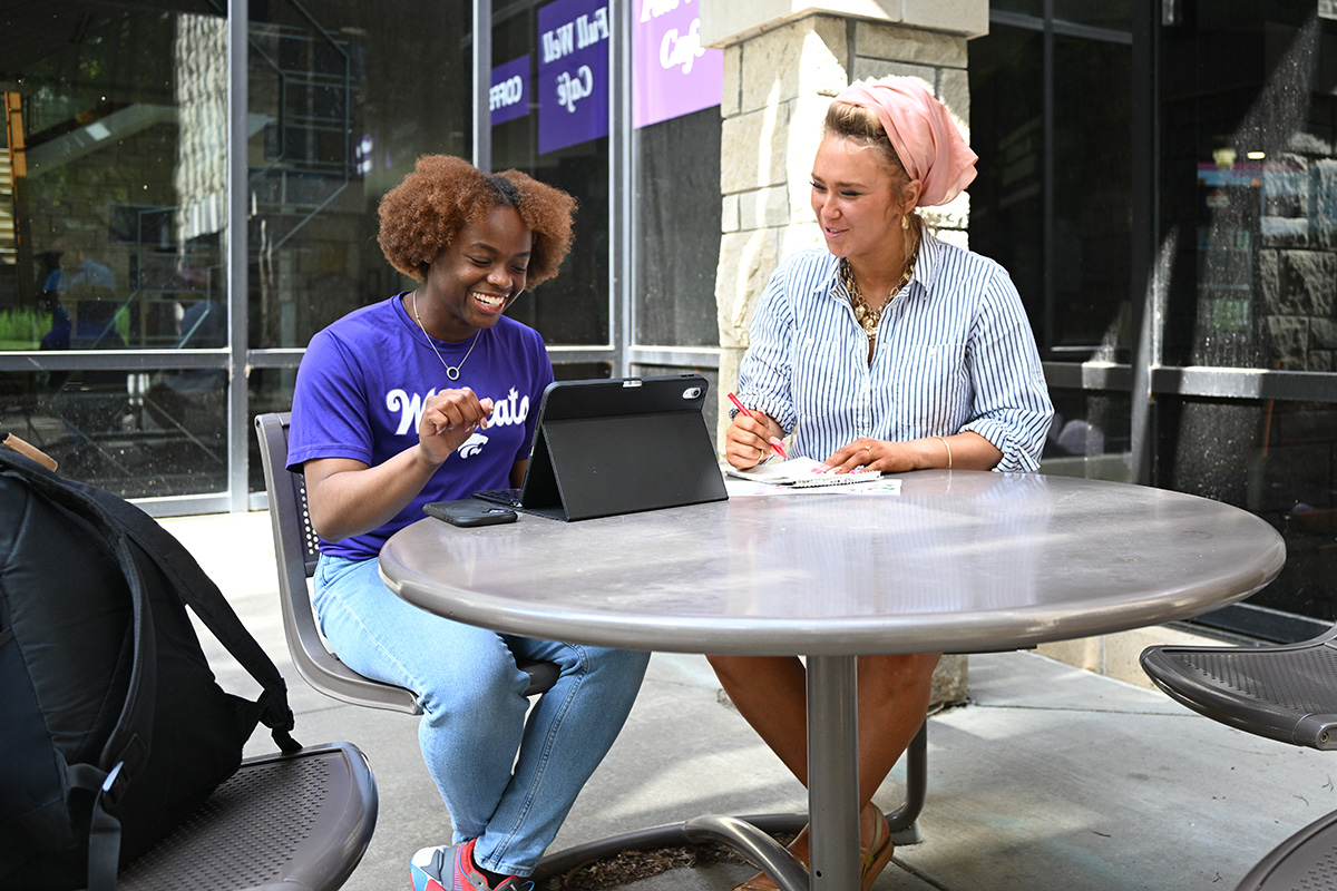 A student in a purple t-shirt with an ipad sits next to a woman wearing a striped shirt and a head wrap at an outdoor table.