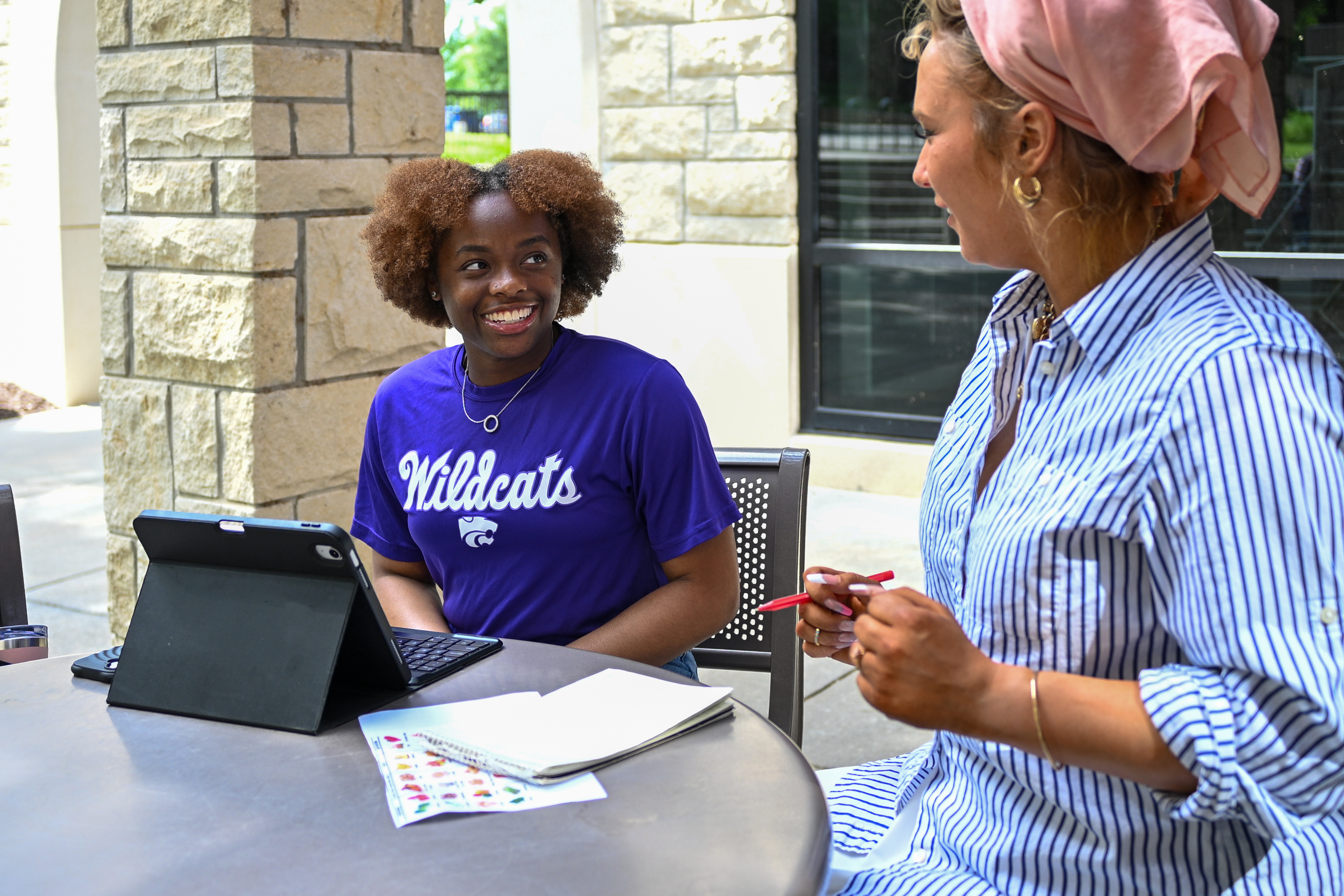 A student in a purple t-shirt with an ipad sits next to a woman wearing a striped shirt and a head wrap at an outdoor table.