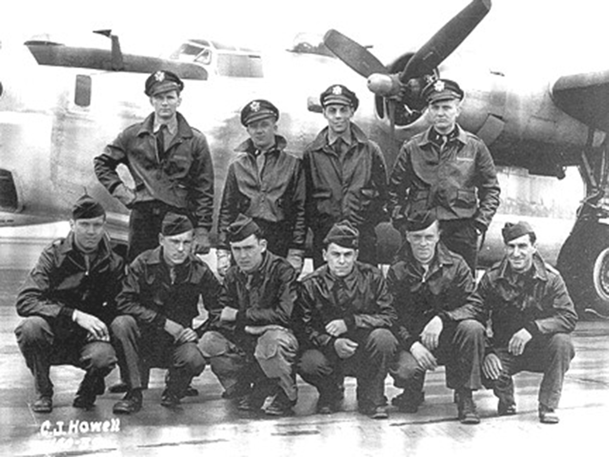 A group of men in military uniforms poses for a photo in front of a large airplane on a sunny day.