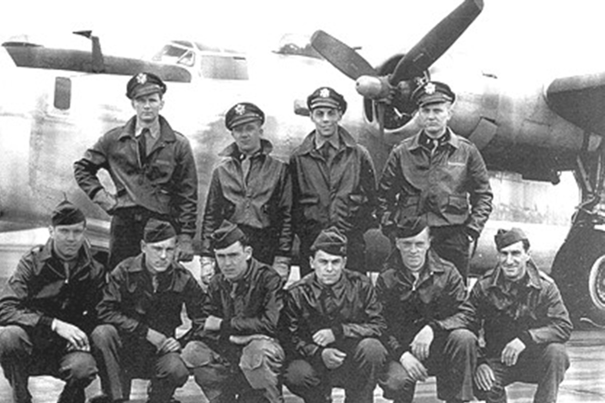 A group of men in military uniforms poses for a photo in front of a large airplane on a sunny day.