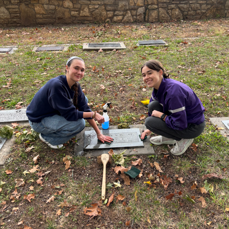 Two college students squat and clean a flat headstone in a cemetery.