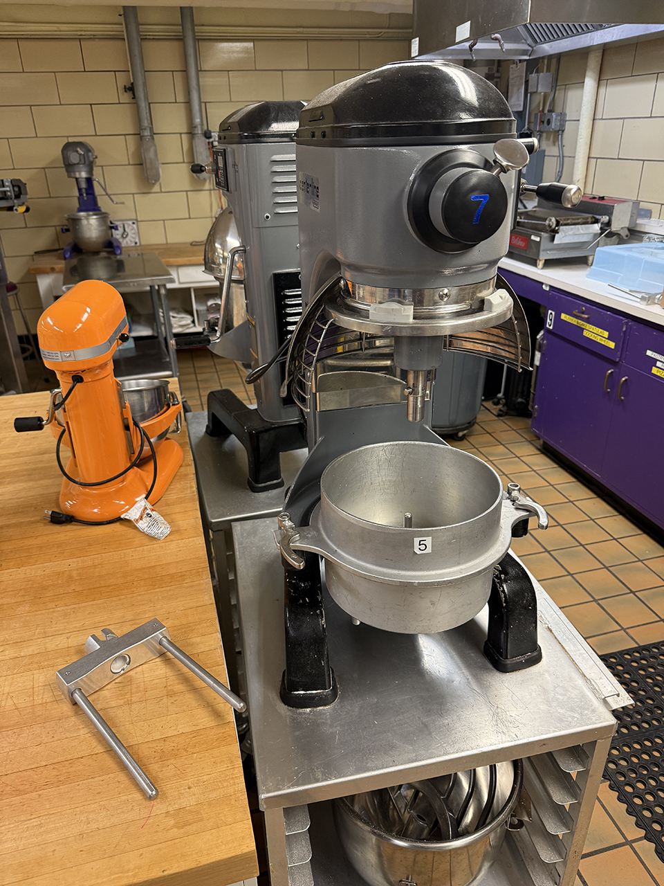 An industrial mixer is shown disassembled, with the mixing attachment laying to the side on a wooden table.