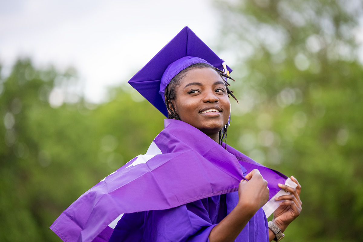 A female college graduate wears a purple cap and gown and drapes a purple flag over her shoulders. 