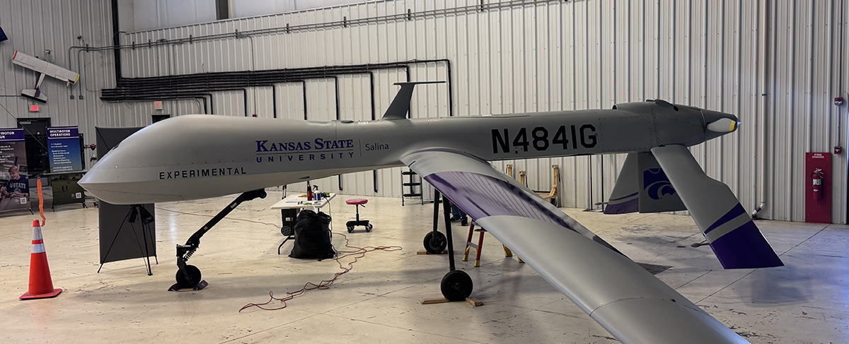 A large gray uncrewed aircraft vehicle with purple K-State Salina wordmark and symbols is in a hangar.