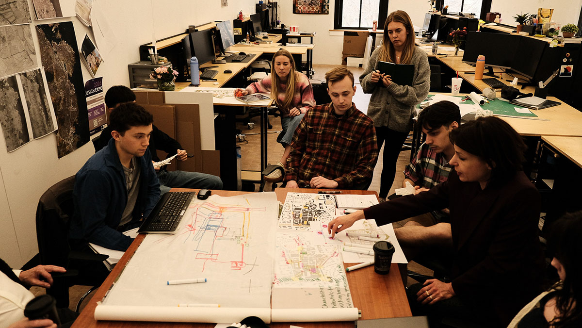 An architecture professor leans over a table and discusses a design document with students.