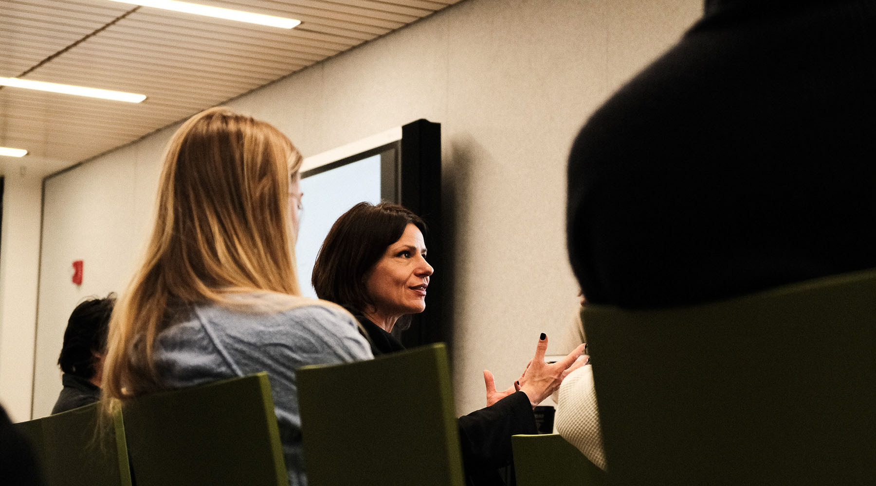 A female college professor discusses architecture while sitting down in a group in a college classroom.