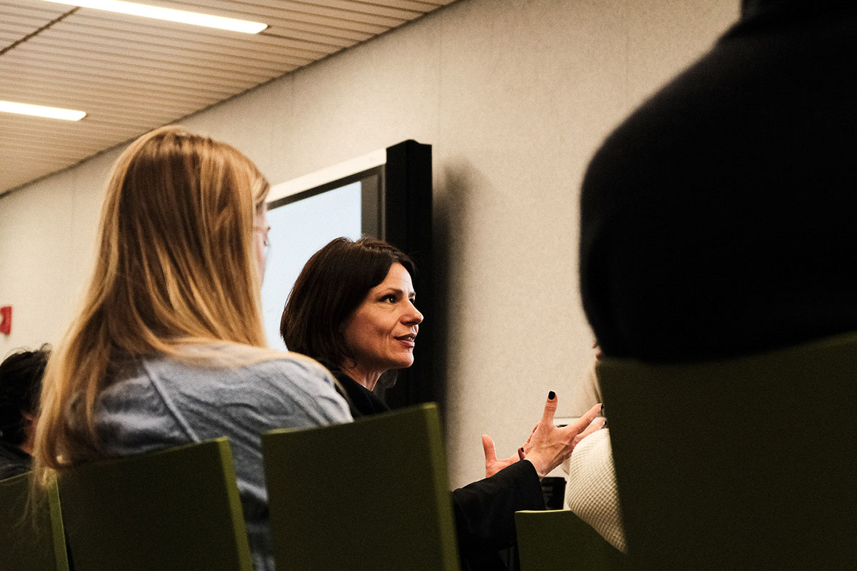 A female college professor discusses architecture while sitting down in a group in a college classroom.