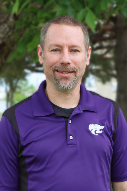 A white man in a purple polo smiles in front of some tree greenery in the background.