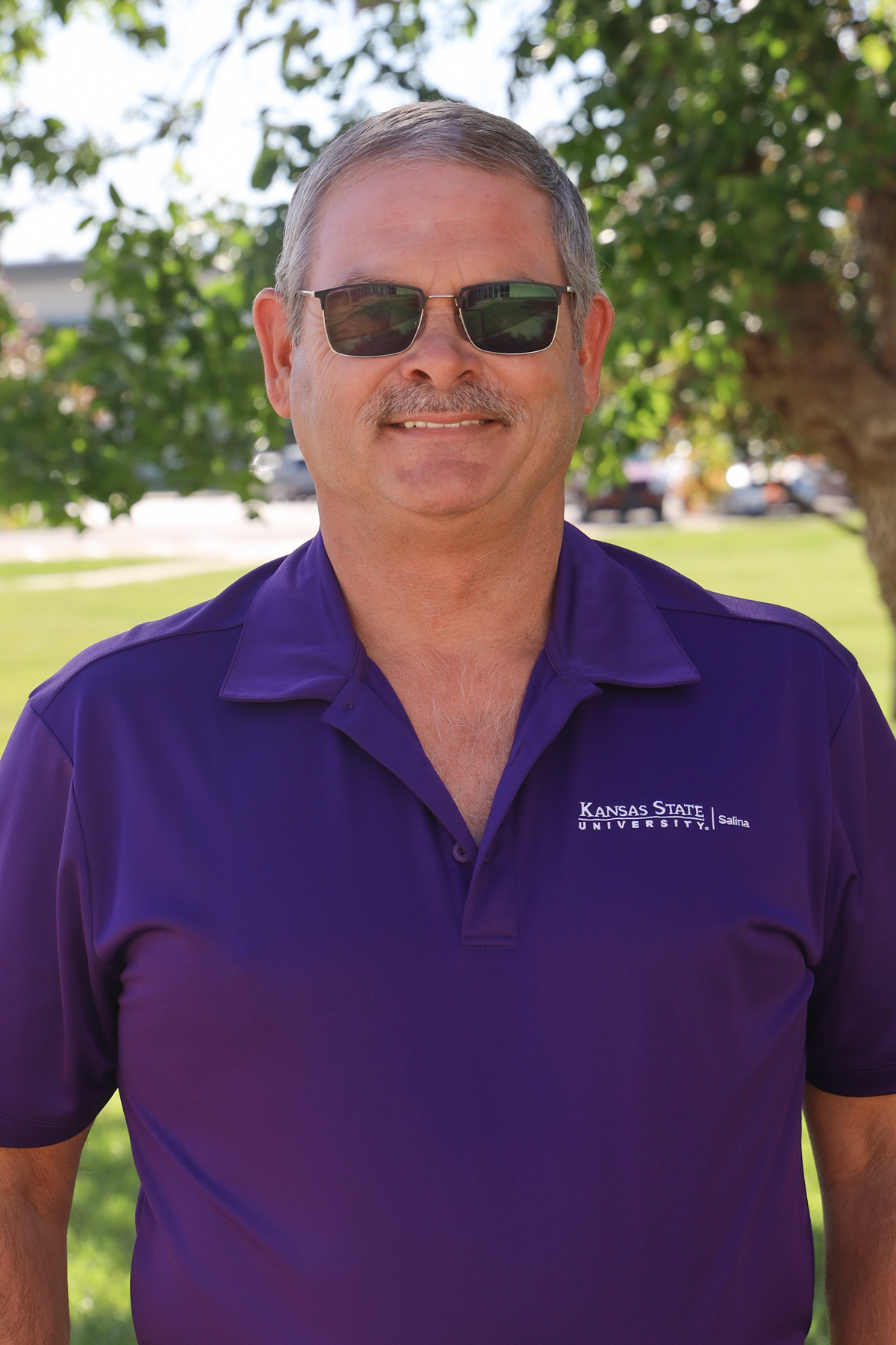 A white man wearing sunglasses and a purple K-State Salina polo smiles with tree greenery in the background.