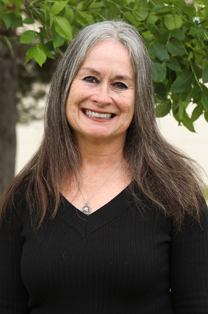 A woman with gray and dark hair wearing a black sweater smiles with tree greenery in the background.